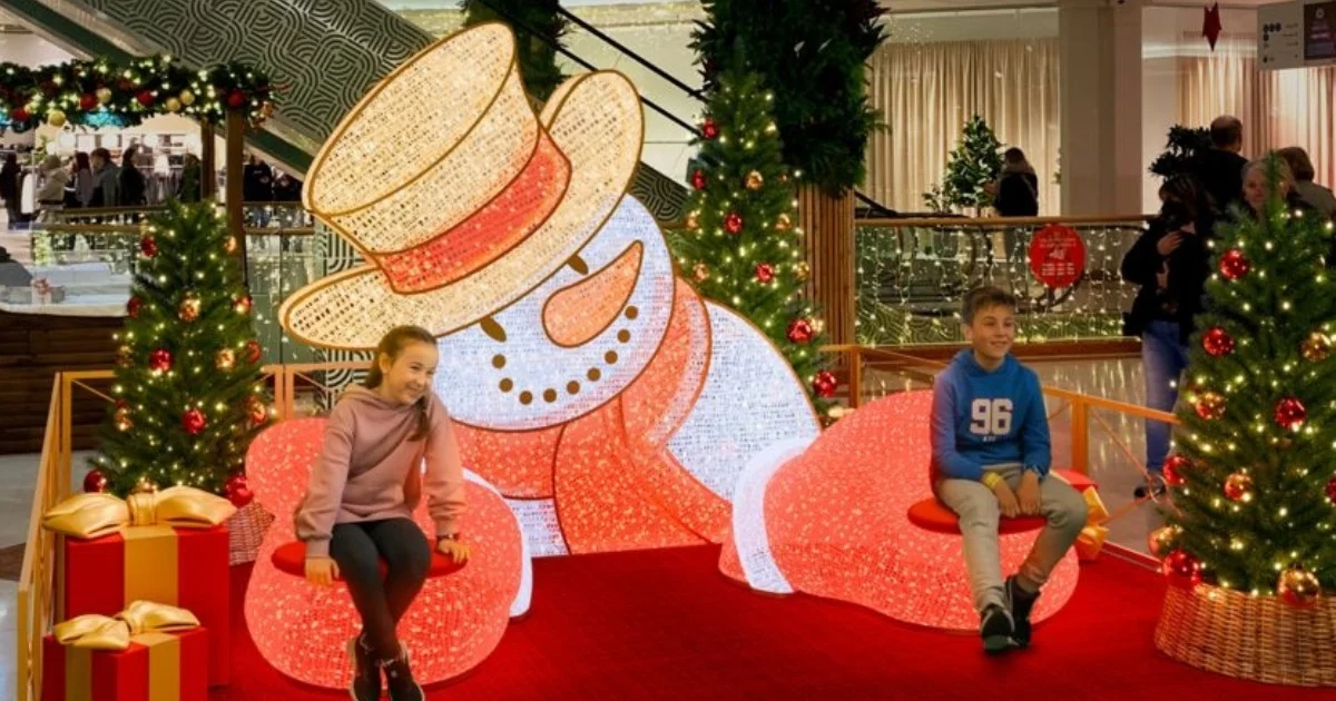 Two children sitting beside a glowing snowman holiday display with decorated Christmas trees and presents in a mall.